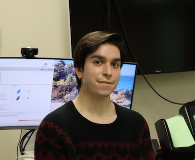 A person with short brown hair wearing a dark sweater with a red and black pattern sits in a research lab. Behind them are computer monitors displaying scientific data and a beach landscape, along with lab equipment. The setting is professional and focused, conveying a calm and studious atmosphere.