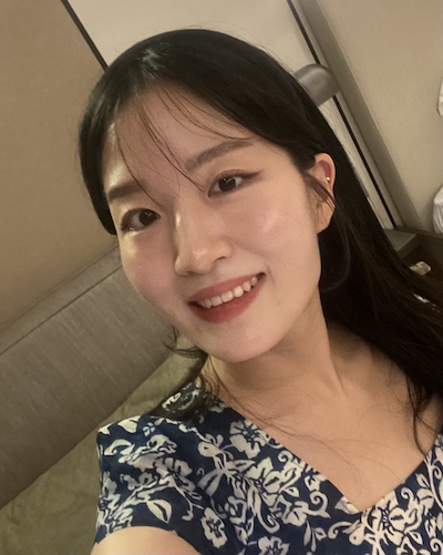 A woman with long black hair wearing a blue dress with white floral patterns sits in a softly lit indoor setting. She is positioned near a beige wall and a cushioned seat, creating a calm and relaxed atmosphere.