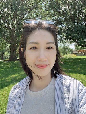 A person with shoulder-length dark hair wearing sunglasses on their head, a light gray t-shirt, and a striped button-up shirt stands outdoors in a sunlit park. The background features green grass, large trees with leafy branches, and a wooden shelter, creating a peaceful and natural atmosphere.